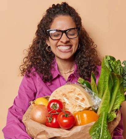 Horizontal shot of happy Brazilian woman with curly hair holds paper bag filled with organic products during her shopping day emphasizes eco friendly attitude to nutrition smiles broadly stands indoor
