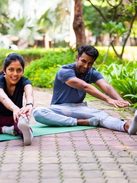 Indian beautiful young couple doing yogasana legs stretching on yoga mat in park outdoor during peaceful morning session focused on flexibility, wellness, balance, holistic healthy lifestyle together